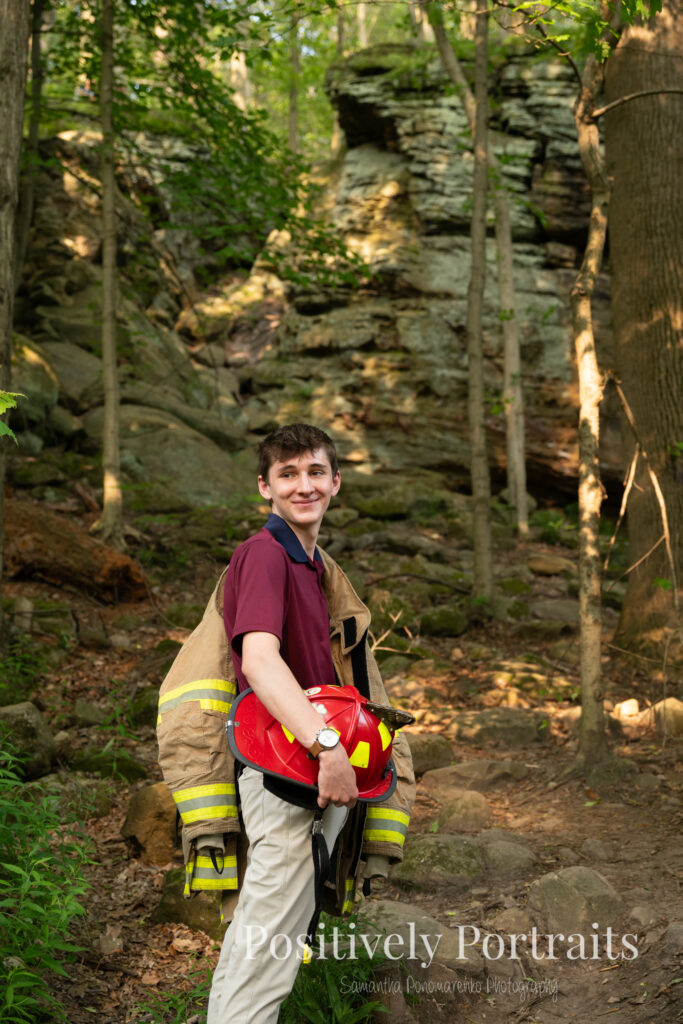 Whipps Ledges Hinckley Ohio Senior Photos Brunswick Ohio. Senior boy wearing firefighter jacket and helmet on rocky ledge at Whipps Ledges with forest backdrop, symbolizing future career and strength