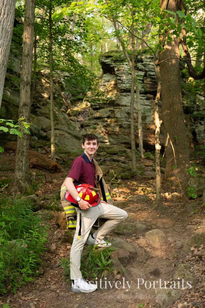 Whipps Ledges Hinckley Ohio Senior Photos Brunswick Ohio. Senior boy wearing firefighter jacket and helmet on rocky ledge at Whipps Ledges with forest backdrop, symbolizing future career and strength