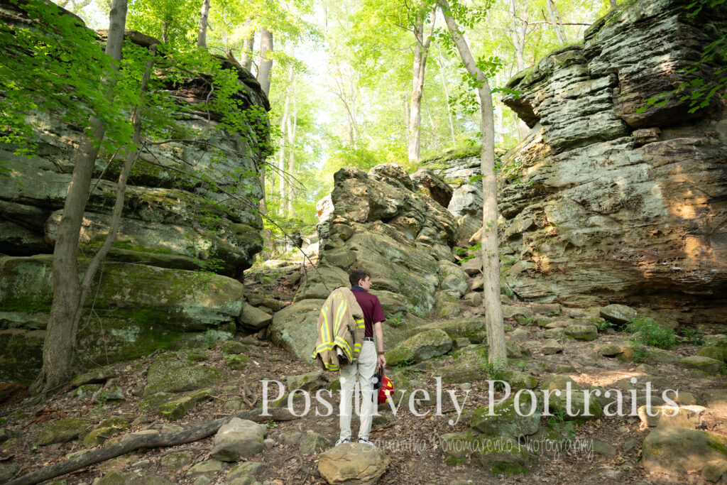 Whipps Ledges Hinckley Ohio Senior Photos Brunswick Ohio. Senior boy wearing firefighter jacket and helmet on rocky ledge at Whipps Ledges with forest backdrop, symbolizing future career and strength