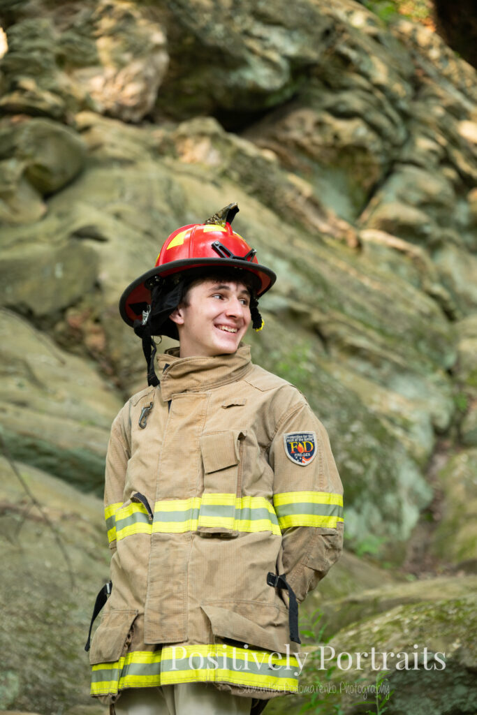Whipps Ledges Hinckley Ohio Senior Photos Brunswick Ohio. Senior boy wearing firefighter jacket and helmet on rocky ledge at Whipps Ledges with forest backdrop, symbolizing future career and strength