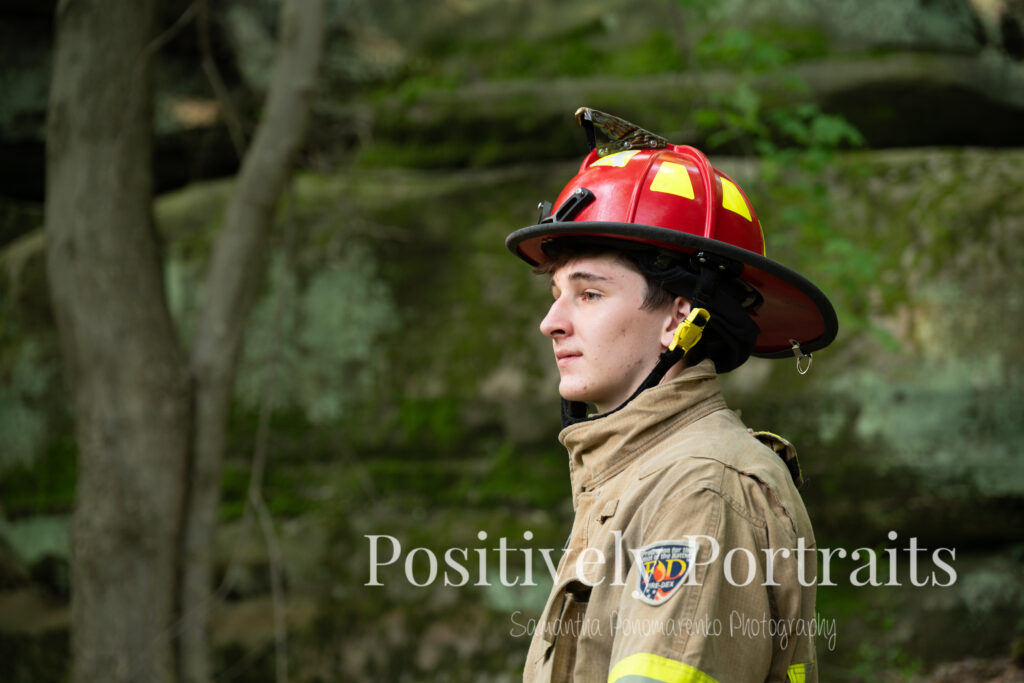 Whipps Ledges Hinckley Ohio Senior Photos Brunswick Ohio. Senior boy wearing firefighter jacket and helmet on rocky ledge at Whipps Ledges with forest backdrop, symbolizing future career and strength