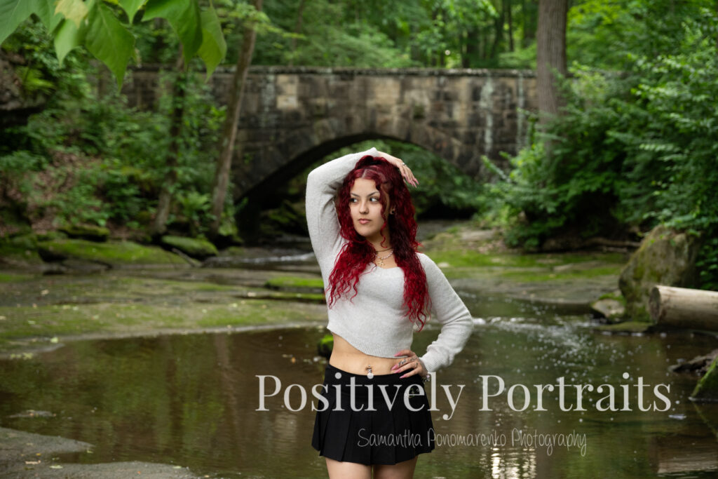 Jocelyn poses confidently on a large rock above the Rocky River at David Fortier River Park in Olmsted Falls, Ohio, wearing a black skirt and gray sweater, surrounded by flowing water and lush greenery. Her senior portraits capture both elegance and adventure, with additional images highlighting stone textures, ivy-covered walls, and classic architecture. The session includes a bold look with a black dress and leopard heels, and ends with cap and gown photos by the riverside, celebrating her graduation.