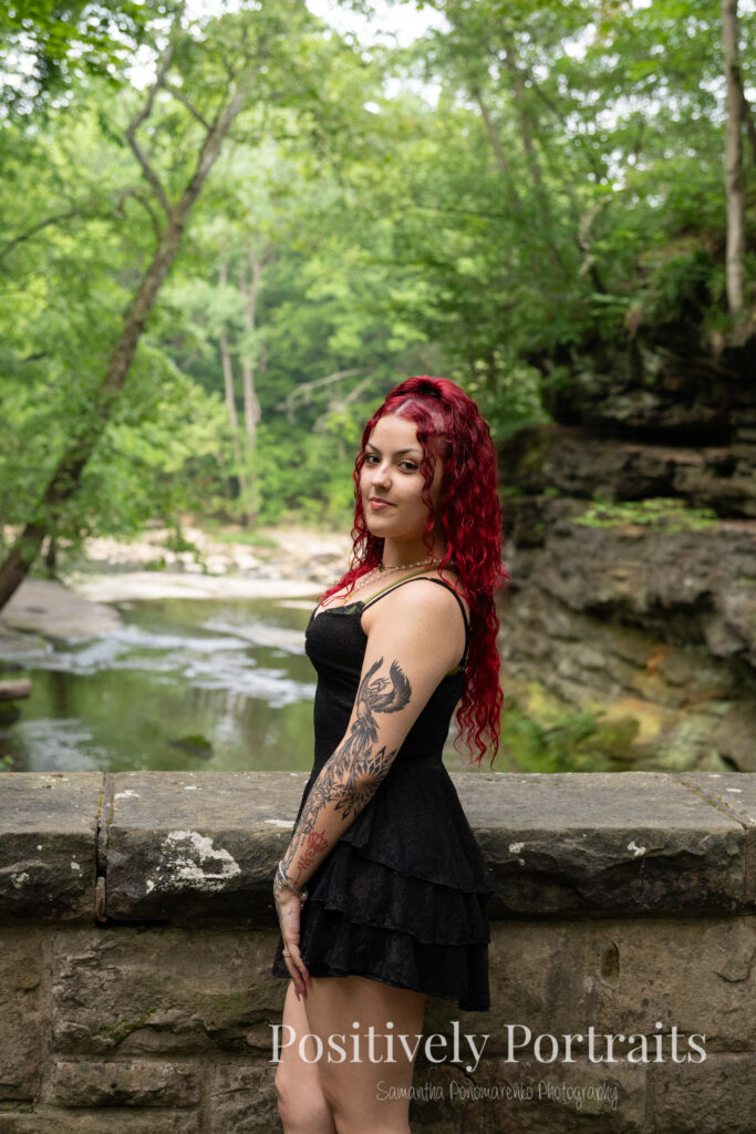 Jocelyn poses confidently near the Rocky River at David Fortier River Park in Olmsted Falls, Ohio by flowing water and lush greenery. Her senior portraits capture both elegance and adventure, with additional images highlighting stone textures, ivy-covered walls, and classic architecture. The session includes a bold look with a black dress and leopard heels, and ends with cap and gown photos by the riverside, celebrating her graduation.