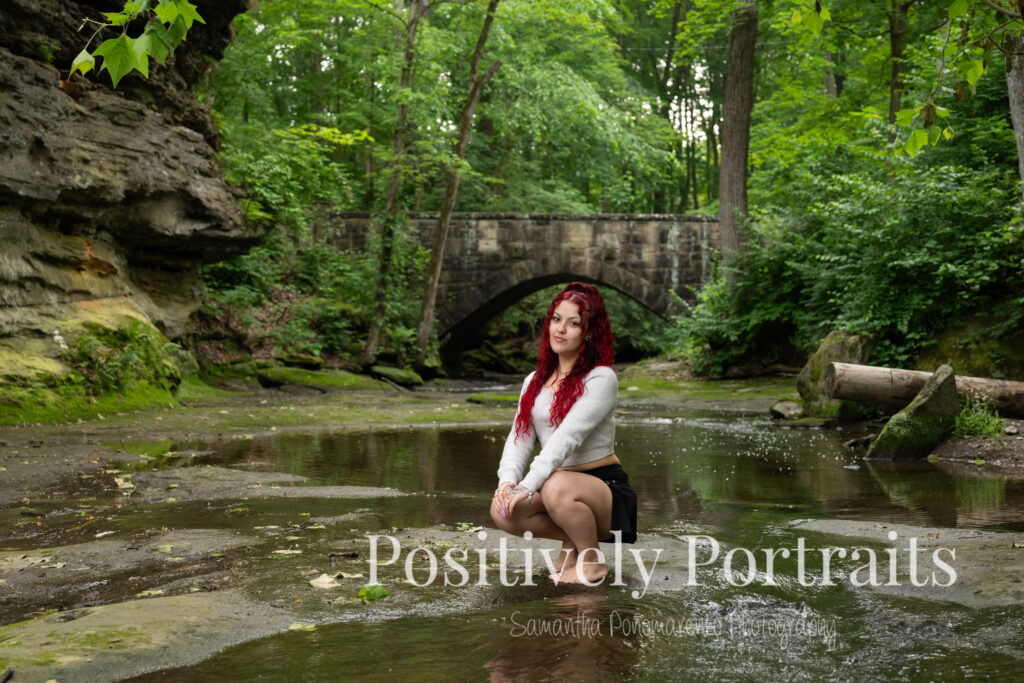 Jocelyn poses confidently on a large rock above the Rocky River at David Fortier River Park in Olmsted Falls, Ohio, wearing a black skirt and gray sweater, surrounded by flowing water and lush greenery. Her senior portraits capture both elegance and adventure, with additional images highlighting stone textures, ivy-covered walls, and classic architecture. The session includes a bold look with a black dress and leopard heels, and ends with cap and gown photos by the riverside, celebrating her graduation.