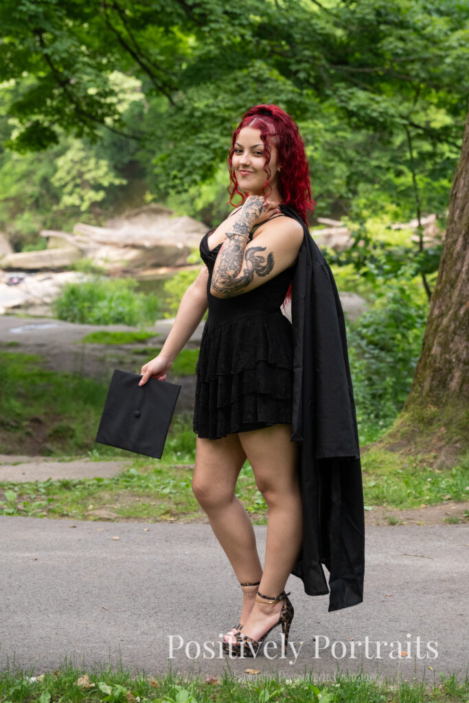 Jocelyn poses confidently near the Rocky River at David Fortier River Park in Olmsted Falls, Ohio by flowing water and lush greenery. Her senior portraits capture both elegance and adventure, with additional images highlighting stone textures, ivy-covered walls, and classic architecture. The session includes a bold look with a black dress and leopard heels, and ends with cap and gown photos by the riverside, celebrating her graduation.