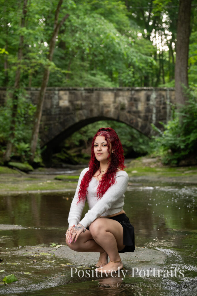 Jocelyn poses confidently on a large rock above the Rocky River at David Fortier River Park in Olmsted Falls, Ohio, wearing a black skirt and gray sweater, surrounded by flowing water and lush greenery. Her senior portraits capture both elegance and adventure, with additional images highlighting stone textures, ivy-covered walls, and classic architecture. The session includes a bold look with a black dress and leopard heels, and ends with cap and gown photos by the riverside, celebrating her graduation.