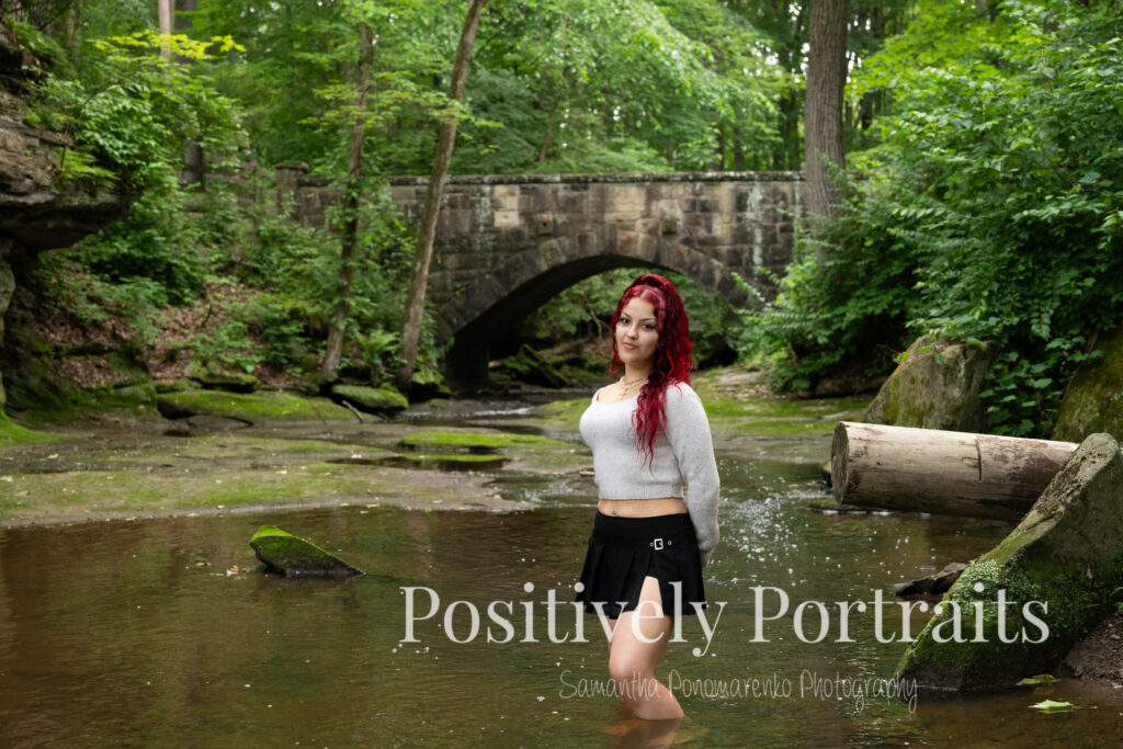 Jocelyn poses confidently on a large rock above the Rocky River at David Fortier River Park in Olmsted Falls, Ohio, wearing a black skirt and gray sweater, surrounded by flowing water and lush greenery. Her senior portraits capture both elegance and adventure, with additional images highlighting stone textures, ivy-covered walls, and classic architecture. The session includes a bold look with a black dress and leopard heels, and ends with cap and gown photos by the riverside, celebrating her graduation.