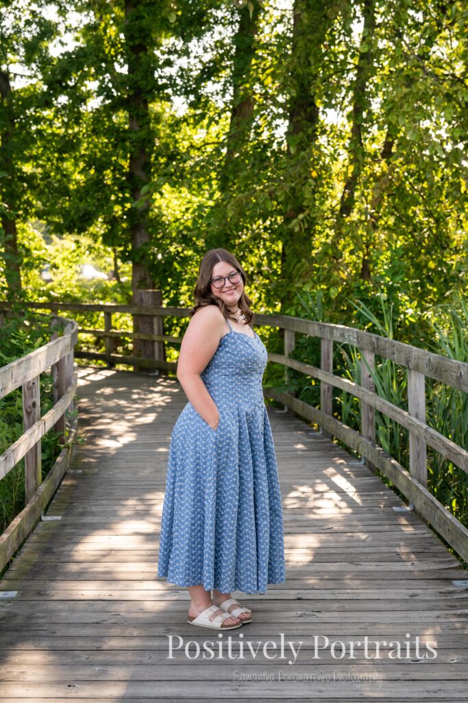 Tabitha in a blue floral midi dress walking across the wooden bridge at Brunswick Lake during her senior photo session, surrounded by soft natural light and calm water in the background.