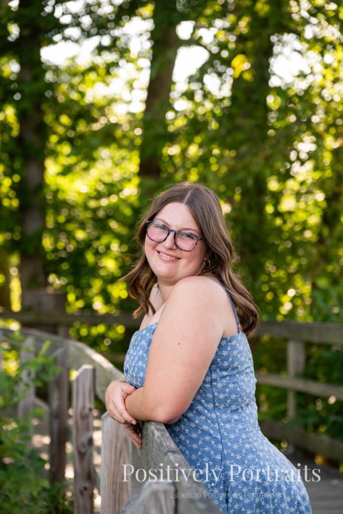 Tabitha in a blue floral midi dress walking across the wooden bridge at Brunswick Lake during her senior photo session, surrounded by soft natural light and calm water in the background.