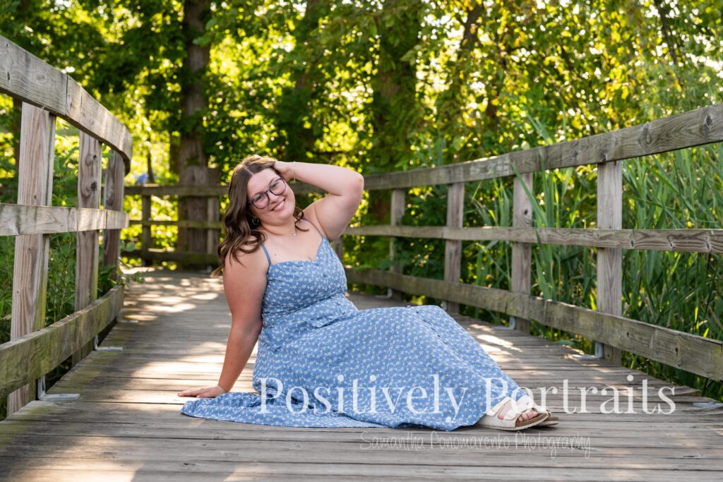 Tabitha in a blue floral midi dress walking across the wooden bridge at Brunswick Lake during her senior photo session, surrounded by soft natural light and calm water in the background.