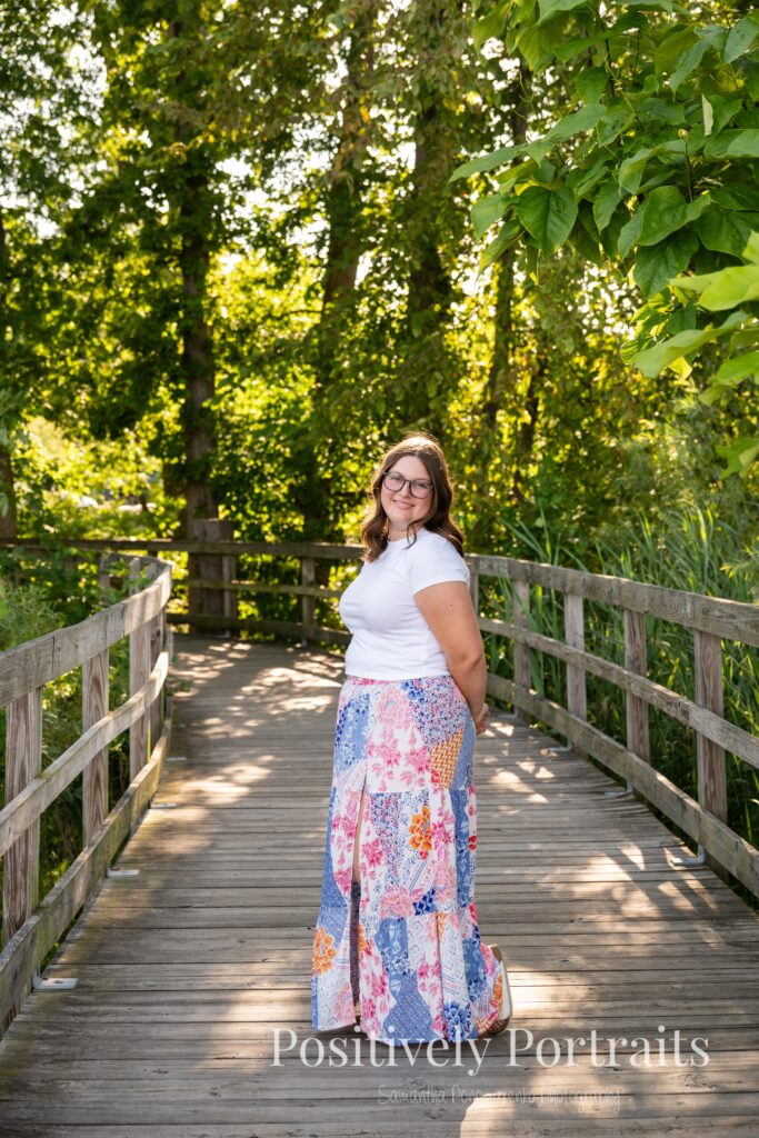 Tabitha in a white t-shirt and colorful patchwork skirt standing on a wooded trail at Brunswick Lake, her outfit’s vibrant patterns contrasting with the greenery.