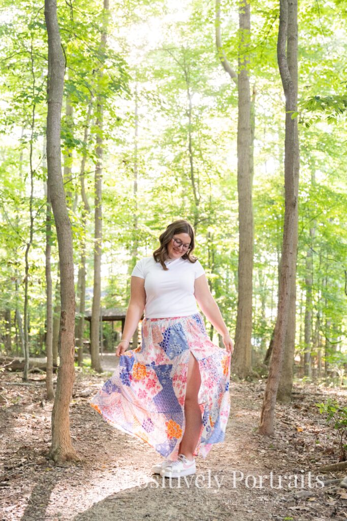 Tabitha in a white t-shirt and colorful patchwork skirt standing on a wooded trail at Brunswick Lake, her outfit’s vibrant patterns contrasting with the greenery.