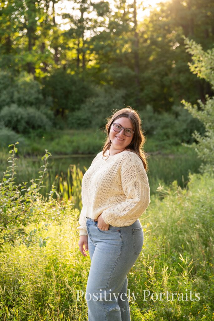 Tabitha standing by the lake’s edge at Brunswick Lake with soft ripples in the water, wearing a yellow sweater and jeans for her senior photos.