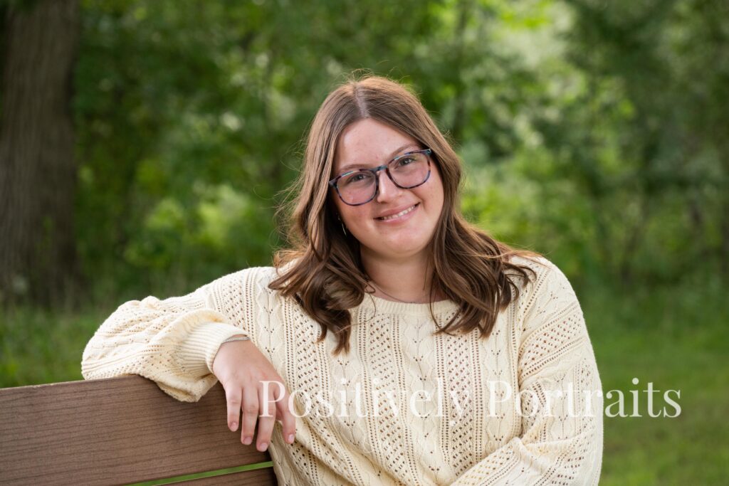 Tabitha standing by the lake’s edge at Brunswick Lake with soft ripples in the water, wearing a yellow sweater and jeans for her senior photos.