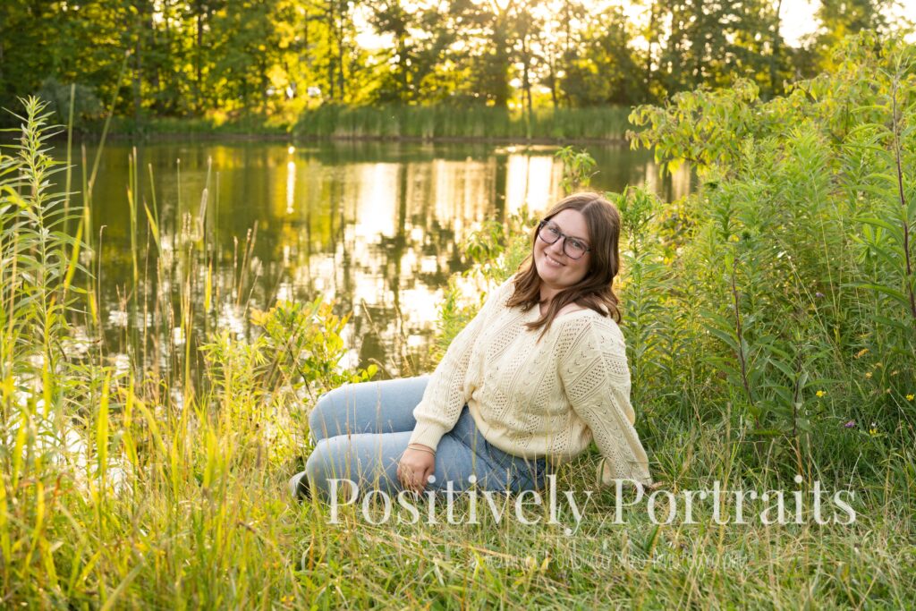 Tabitha standing by the lake’s edge at Brunswick Lake with soft ripples in the water, wearing a yellow sweater and jeans for her senior photos.